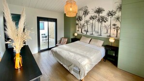 Une chambre à coucher comprenant un lit, une table de chevet, une lampe, une chaise, un vase contenant de l’herbe des pampas et une fresque représentant des palmiers.