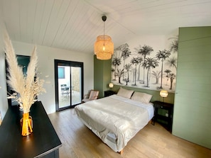 Une chambre à coucher comprenant un lit, une table de chevet, une lampe, une chaise, un vase contenant de l’herbe des pampas et une fresque représentant des palmiers.