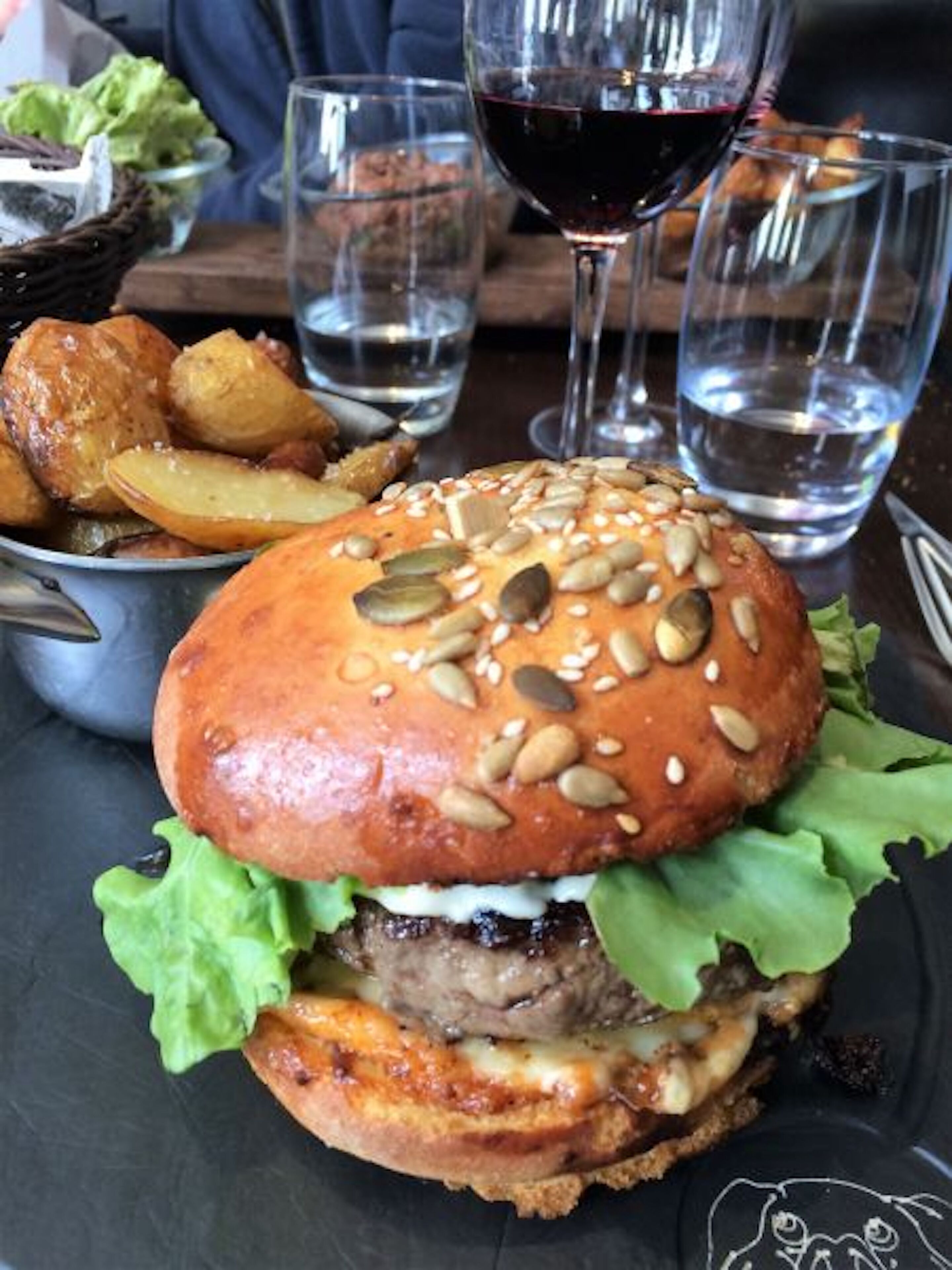 Ein Burger mit Sesambrötchen, Salat, Käse und einem Rinderpatty, zusammen mit Pommes und einem Glas Rotwein auf einem Tisch.