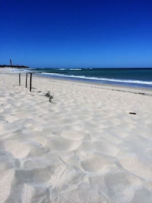 Une plage avec un ciel dégagé, un rivage sablonneux et un phare à l’horizon.