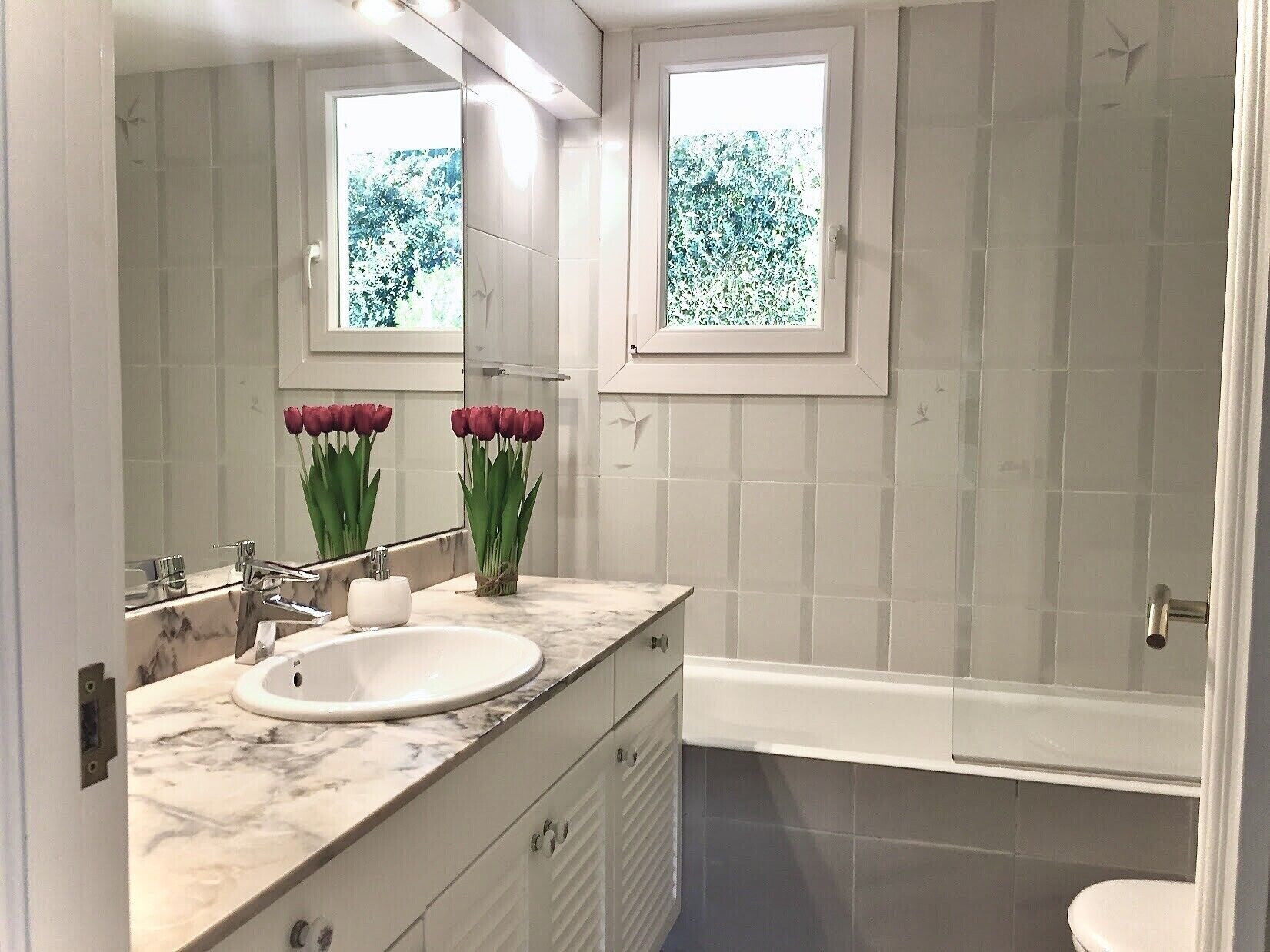 Un baño moderno con tocador de mármol, un espejo grande y una ventana con vistas a zonas verdes.