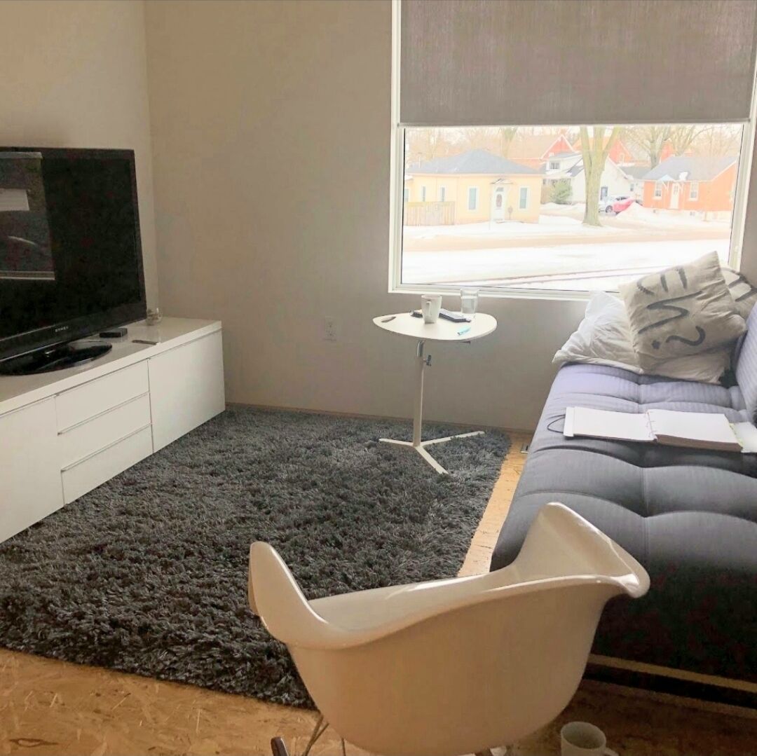 A modern living room with a flat-screen TV on a white cabinet, a black shag carpet, a white chair, a small white table, and a sofa with a window view.