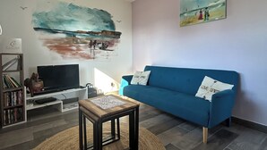 Una sala de estar con un sofá azul, un televisor sobre un mueble blanco, un cuadro de una playa y una alfombra tejida.
