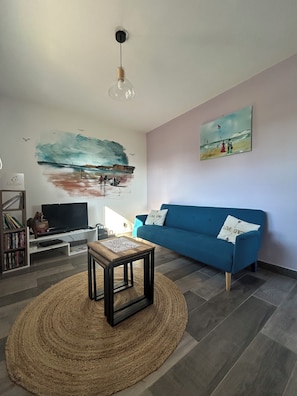 Ett vardagsrum med en blå soffa, en tv på ett vitt tv-bord, en tavla som föreställer en strandscen och en vävd matta.