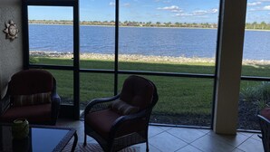 Una habitación con un ventanal grande con vista a un cuerpo de agua, dos sillas de mimbre y una mesita con una vela.