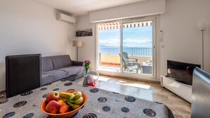 Un salon moderne avec un canapé, un téléviseur, une table à manger ornée de fruits et un balcon offrant une vue sur la mer.