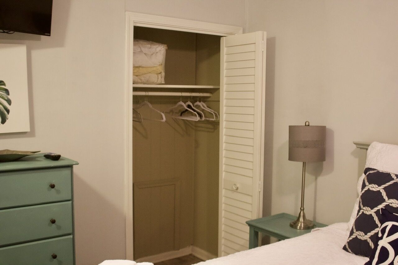 A bedroom with a bed, a nightstand, a bedside table, a lamp, a dresser, and a closet with hangers.