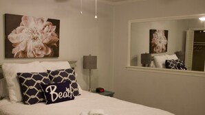 Un lit bien fait, avec une serviette pliée, un oreiller orné du mot «Plage», et un grand tableau floral accroché au-dessus.