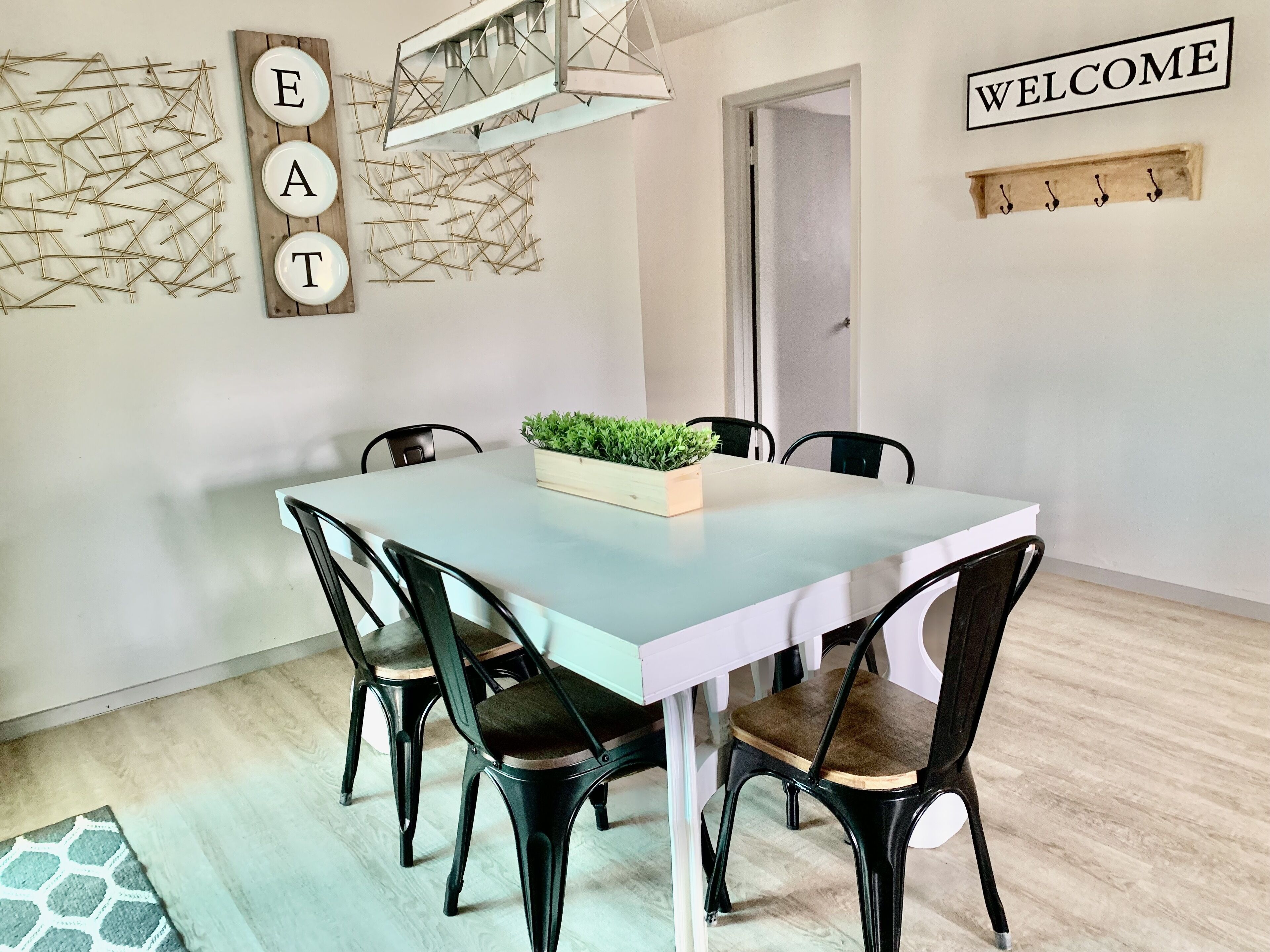 Un coin repas avec une table bleu clair, des chaises noires, une pancarte « BIENVENUE » et une plante fixée au mur.