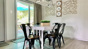 A dining area with a white table, black chairs, and a chandelier. There is a wall decoration with the word "EAT" and a plant on the table.