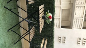 Comedor con mesa y sillas, rodeado por un seto verde y un edificio blanco con contraventanas.