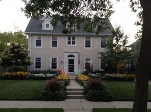 Een tweelaags huis met een veranda, een vlag en een verzorgde tuin met bloemen en struiken.