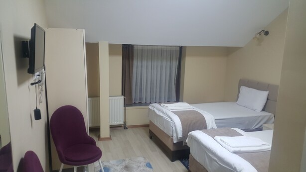 A hotel room with two beds, a desk, a chair, a radiator, and a window with curtains. - Lux Otel (Erzurum)