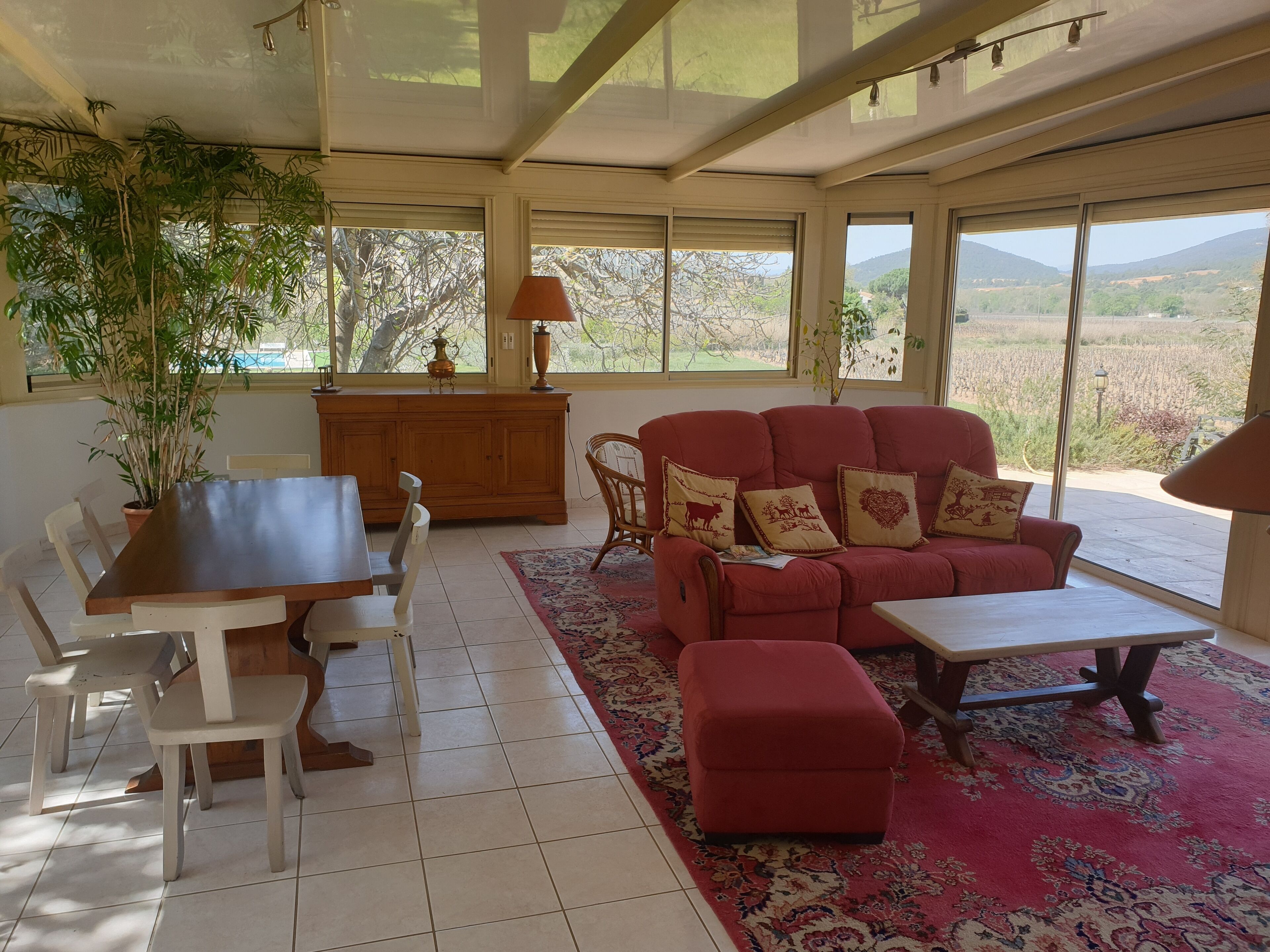 A sunlit room with a dining table, chairs, a sofa with decorative pillows, a rug, and a view of a rural landscape through large windows.
