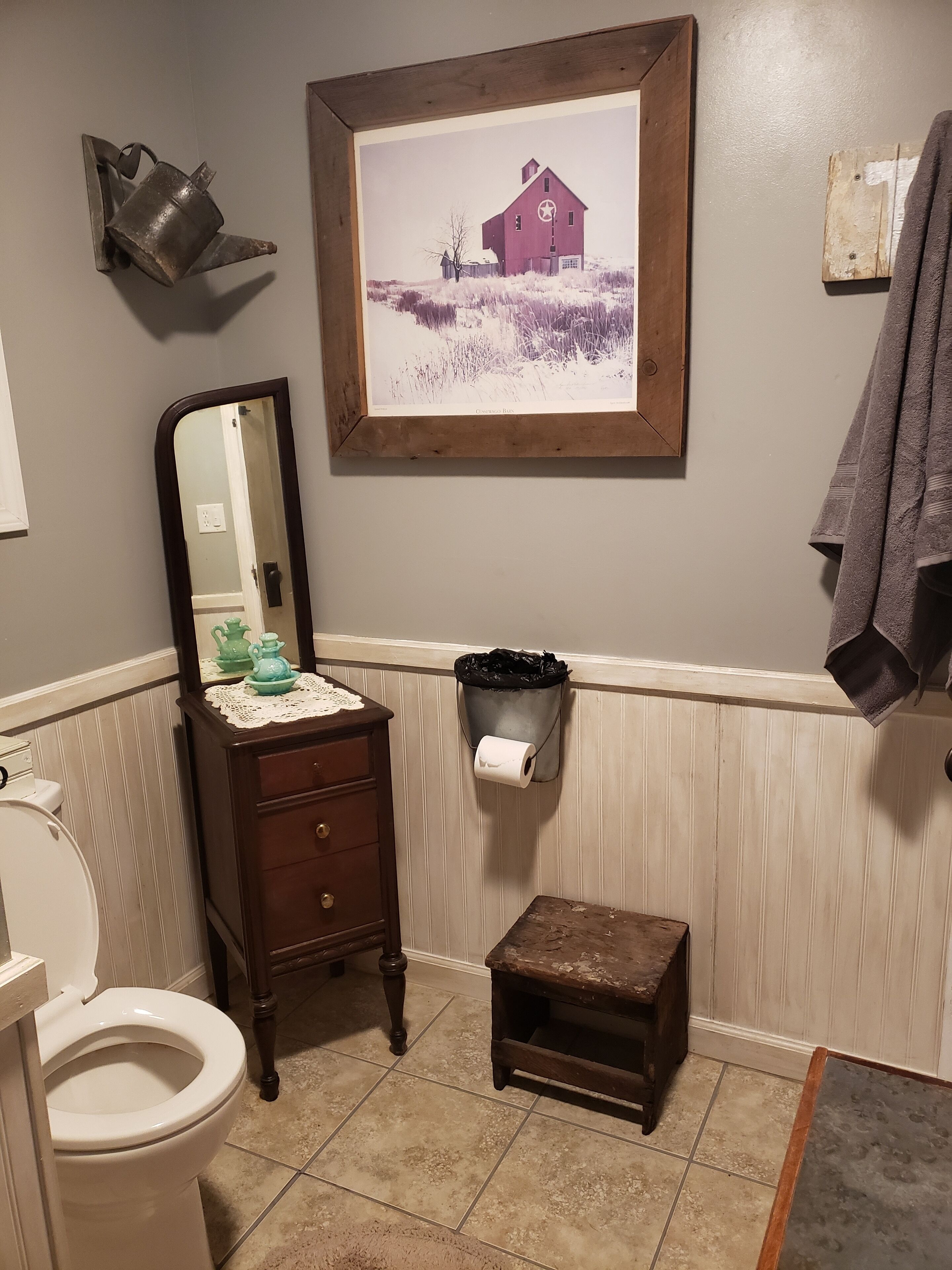 Un baño con inodoro, tocador de madera con espejo, un cuadro con una imagen de un granero y un pequeño cubo de basura.