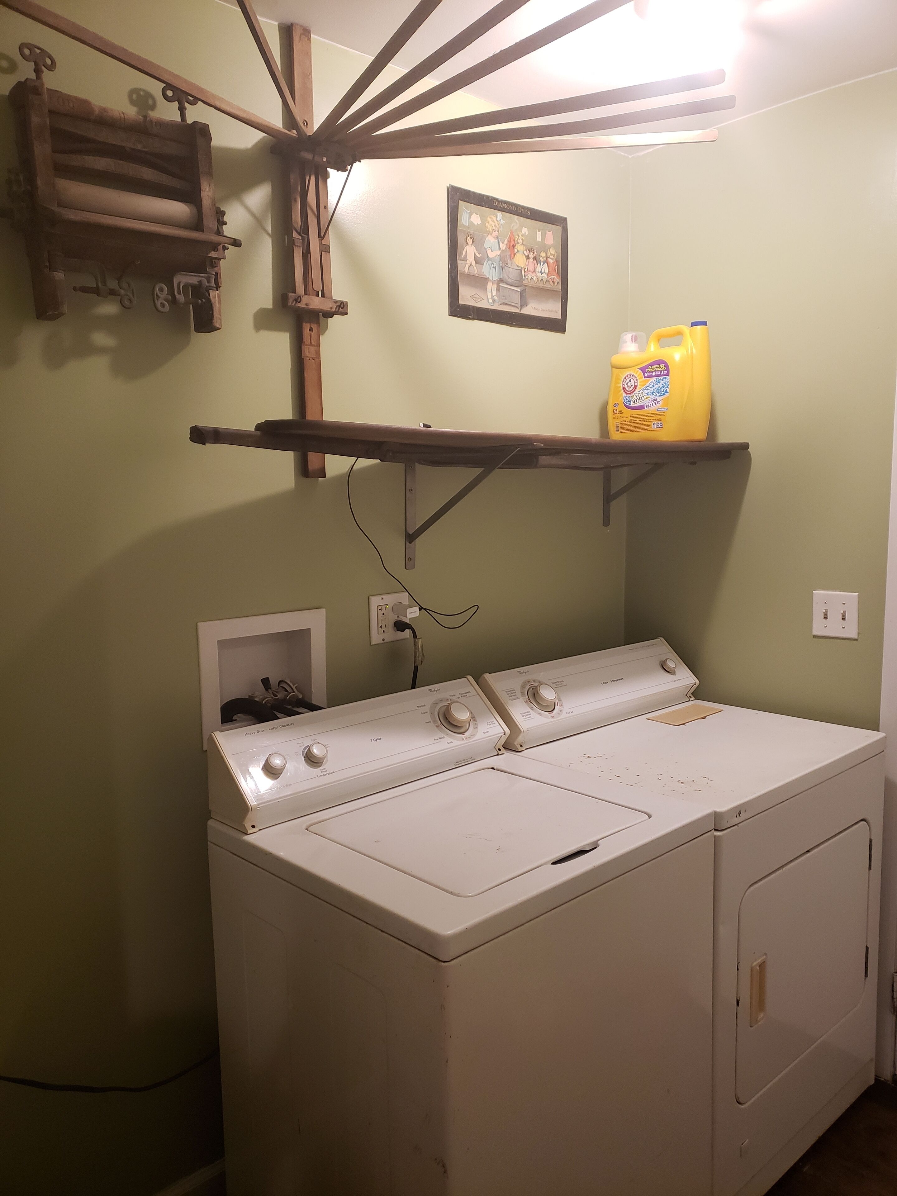 A laundry area with a front-loading washer and dryer, a shelf with a detergent bottle, and a wall-mounted drying rack.