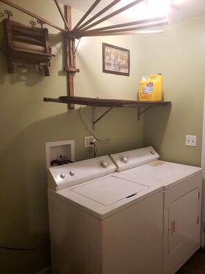A laundry area with a front-loading washer and dryer, a shelf with a detergent bottle, and a wall-mounted drying rack.