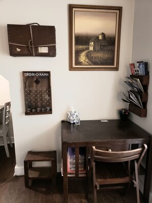 Un coin repas avec une table et des chaises en bois, un tableau encadré au-dessus, un tableau d’affichage avec une affiche vantant les mérites de diverses boissons, et une bibliothèque remplie de magazines.