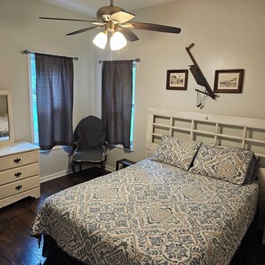 Une chambre à coucher comprenant un lit, une chaise, une commode, un ventilateur de plafond et deux tableaux encadrés accrochés au mur.