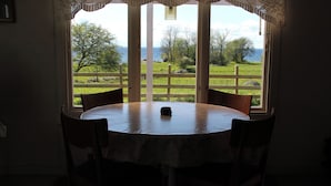 Mesa redonda con mantel blanco y cuatro sillas, ubicada frente a un gran ventanal con vistas a un paisaje cubierto de césped y árboles.