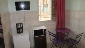 Habitación pequeña con refrigerador blanco, microondas, una mesa con sillas moradas, televisor montado en la pared y ventana con barrotes y cortina roja.