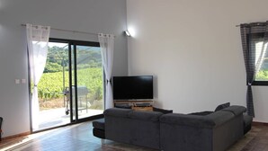 A modern living room with a large grey sofa, a flat-screen TV, and a sliding glass door leading to a patio with a view of a vineyard. - Villa located in the heart of Provence with private pool on 3 hectares. (Roquevaire)