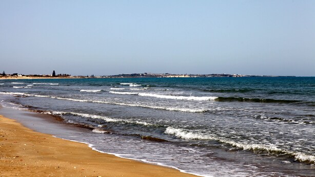 A beach with waves, a clear sky, and a distant shoreline with buildings. - Villa Concita Sicily (viale malta)