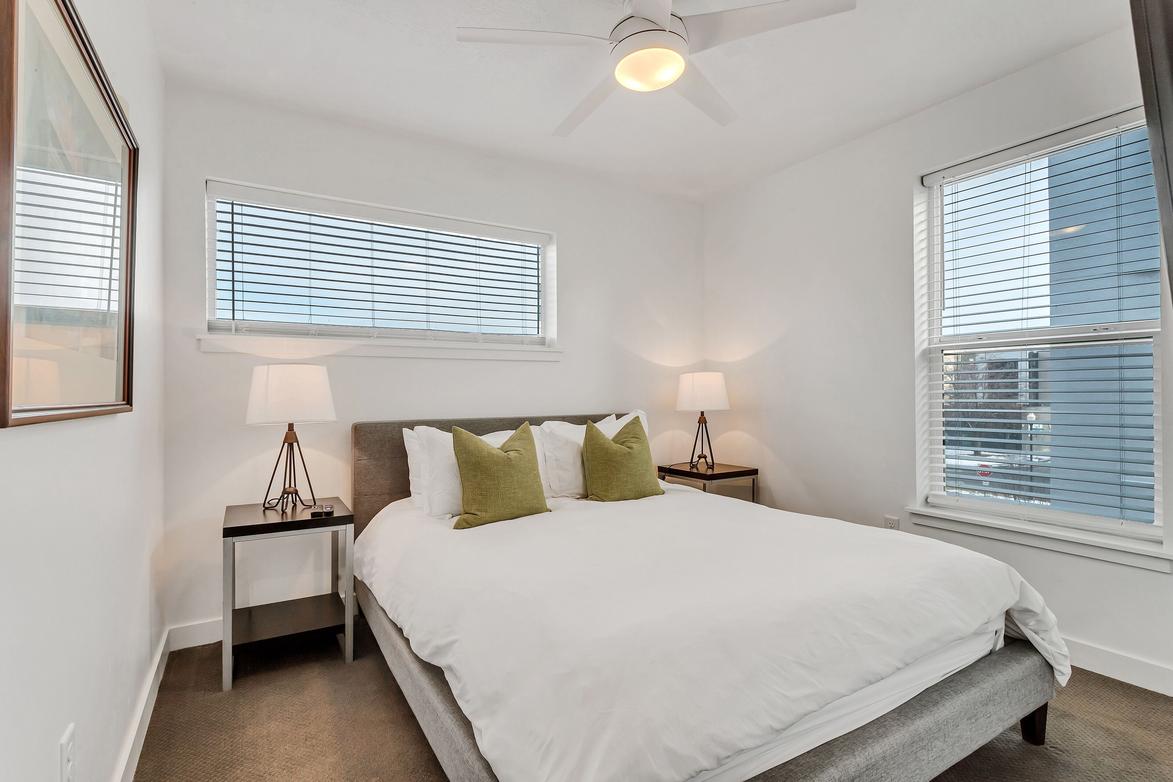 A bedroom with a bed, two nightstands, two lamps, a ceiling fan, and two windows with blinds.