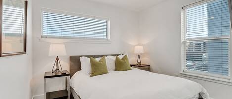 A bedroom with a bed, two nightstands, two lamps, a ceiling fan, and two windows with blinds.