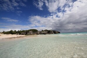 A beach with clear turquoise water, a sandy shore, and a rocky outcrop with greenery.