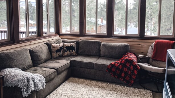 A cozy living room with a sectional sofa, a red plaid throw blanket, and a view of a snowy landscape through large windows. - Beautiful OHV Friendly Lodge, Sun Room, Close Walk to River, Deck, Fire Pit (South Fork)