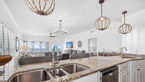 A modern kitchen with granite countertops, stainless steel sinks, and a dishwasher. There are pendant lights, a chandelier, and a view of the living area. - Grand Playa 301, Scenic 30A, Gulf Front Condo, Pool with Private Beach Access (Seagrove Beach)