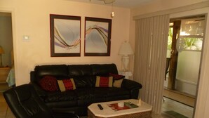 Ein Wohnzimmer mit einer schwarzen Ledersofa, zwei gerahmten abstrakten Gemälden, einem kleinen Holztisch und einem gemusterten Teppich.