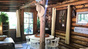 L’intérieur d’un chalet rustique en bois présente une table de salle à manger ronde entourée de chaises blanches, un canapé en osier et un poteau en bois auquel est accroché un chapeau.
