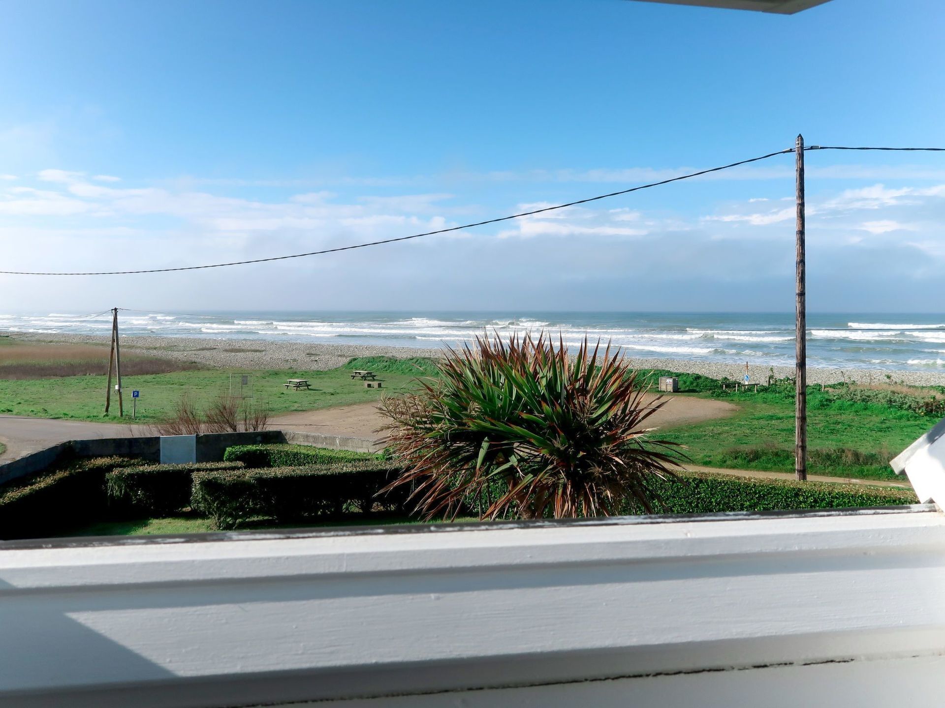 Blick auf den Strand vom Balkon aus, mit blauem Himmel, einer Rasenfläche und einer Holzkonstruktion.