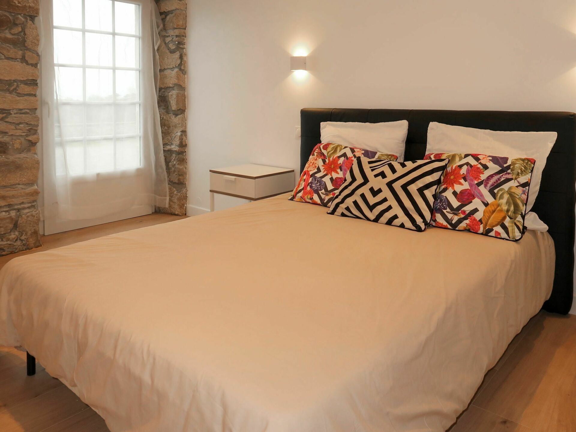 Een net opgemaakt bed met een beige sprei en twee decoratieve kussens, een klein nachtkastje en een wandlamp.