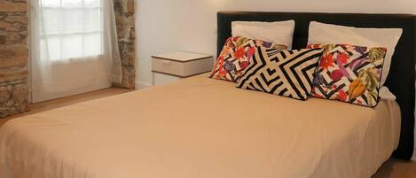 Een net opgemaakt bed met een beige sprei en twee decoratieve kussens, een klein nachtkastje en een wandlamp.