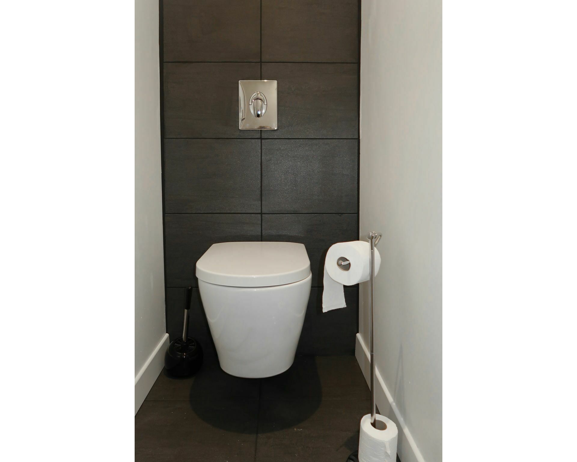 Une petite salle de bain moderne avec des toilettes blanches, des murs recouverts de carreaux foncés et un porte-rouleau de papier toilette.