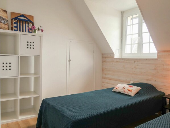 Una stanza accogliente e piccola, con soffitto a falde, un letto singolo con copriletto verde, un armadio bianco con scomparti multipli e una finestra che lascia entrare luce naturale.