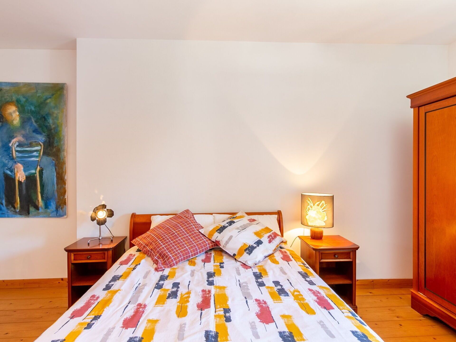 A bedroom with a bed, a nightstand, a lamp, a painting, and a wardrobe.