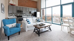 A modern living room with a white sofa, blue armchair, and a coffee table. A kitchen with grey cabinets and a brick wall is visible through large windows.