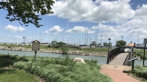 A park with a paved walkway, a sign, and a bridge over a body of water.