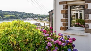 Een kustgezicht met een wit gebouw met stenen details, een raam met uitzicht op een balkon, en weelderige begroeiing op de voorgrond.