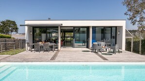 Una casa moderna con piscina, que cuenta con un comedor con mobiliario exterior y un ventanal que da a la zona de la piscina.