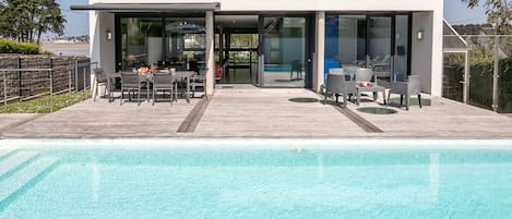 Una casa moderna con piscina, que cuenta con un comedor con mobiliario exterior y un ventanal que da a la zona de la piscina.