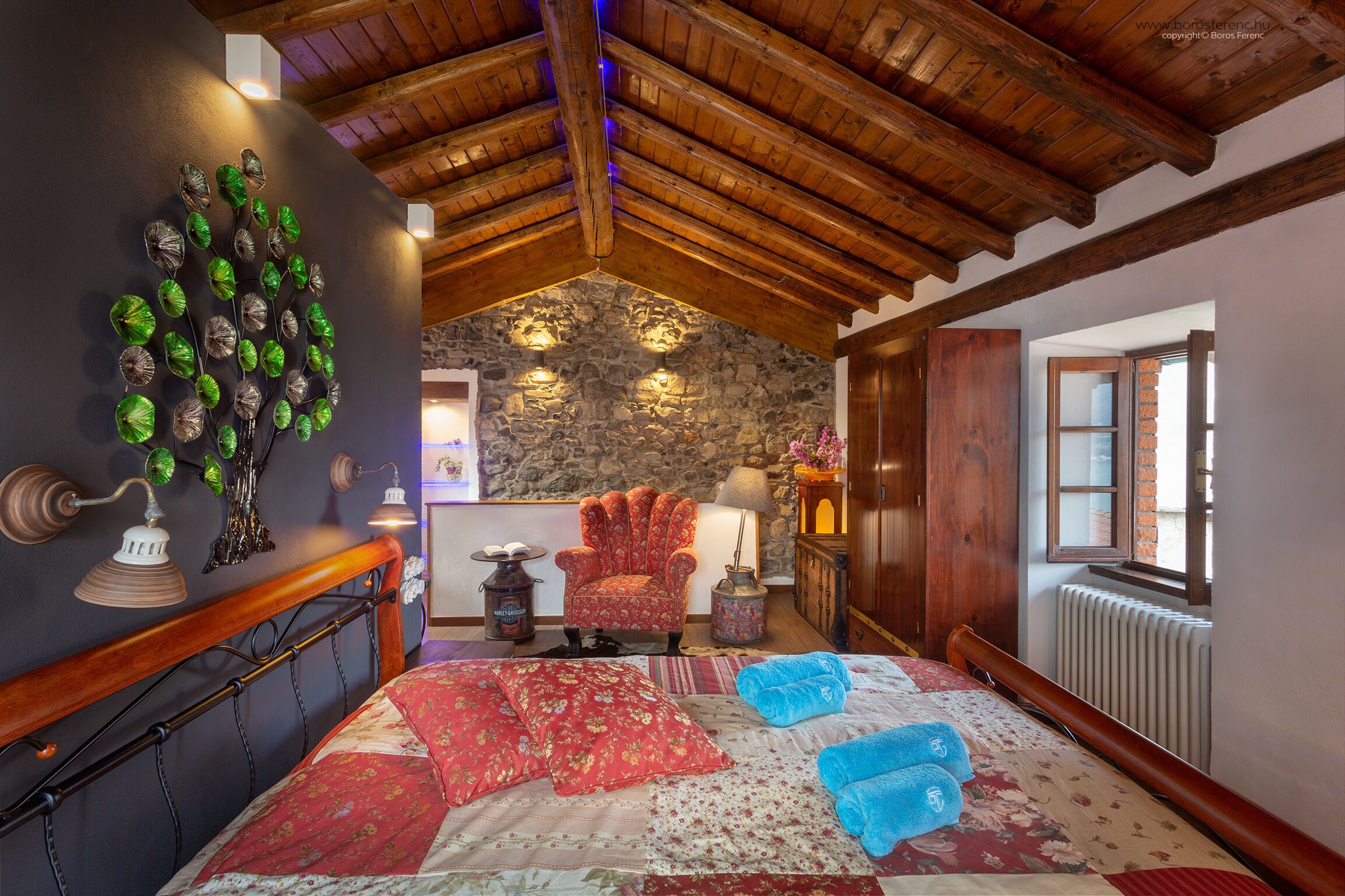 A bedroom with a wooden ceiling, a stone wall, a bed with floral bedding, a red armchair, and a decorative wall piece.