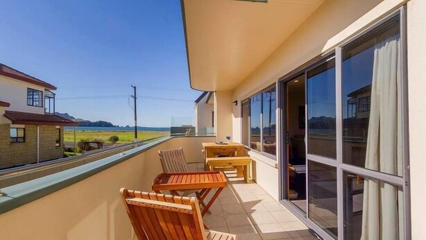 A balcony with wooden chairs and a table, large sliding glass doors, and a view of the sea and hills.