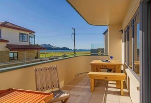 A balcony with wooden furniture, a view of a house and mountains, and a clear sky.