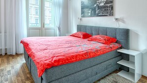 A bedroom with a bed, a nightstand, a window with curtains, and a wall-mounted picture. - ValenciaKV Apartments (Karlovy Vary)