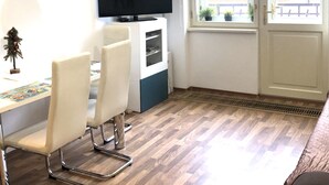 A small, bright room with a wooden floor, a white desk with a computer, a television, and a window with a view of buildings. - ValenciaKV Apartments (Karlovy Vary)